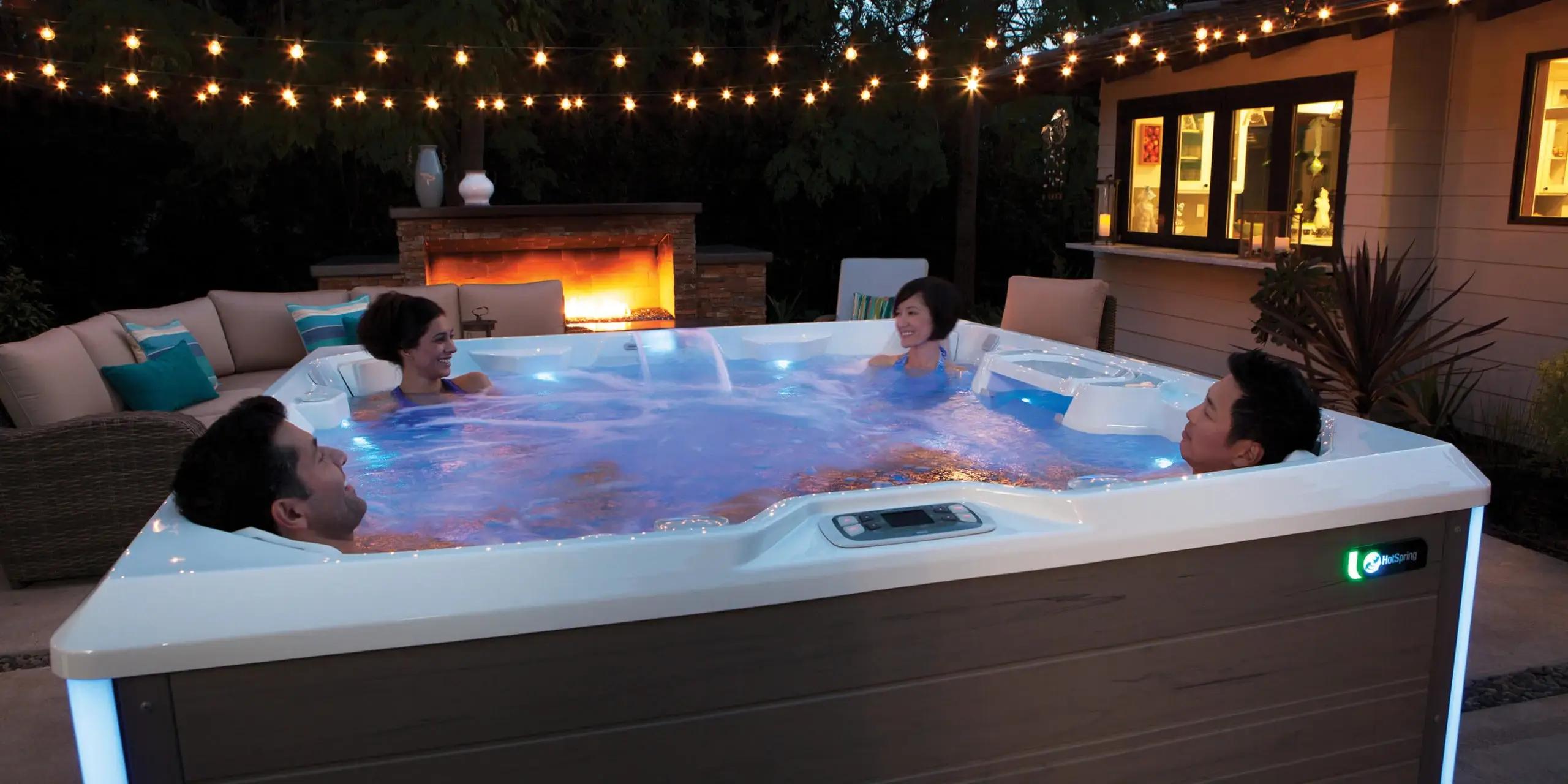 Four people in a Limelight&reg; spa after dark, the water lit blue, with string lights overhead and a fire feature glowing behind