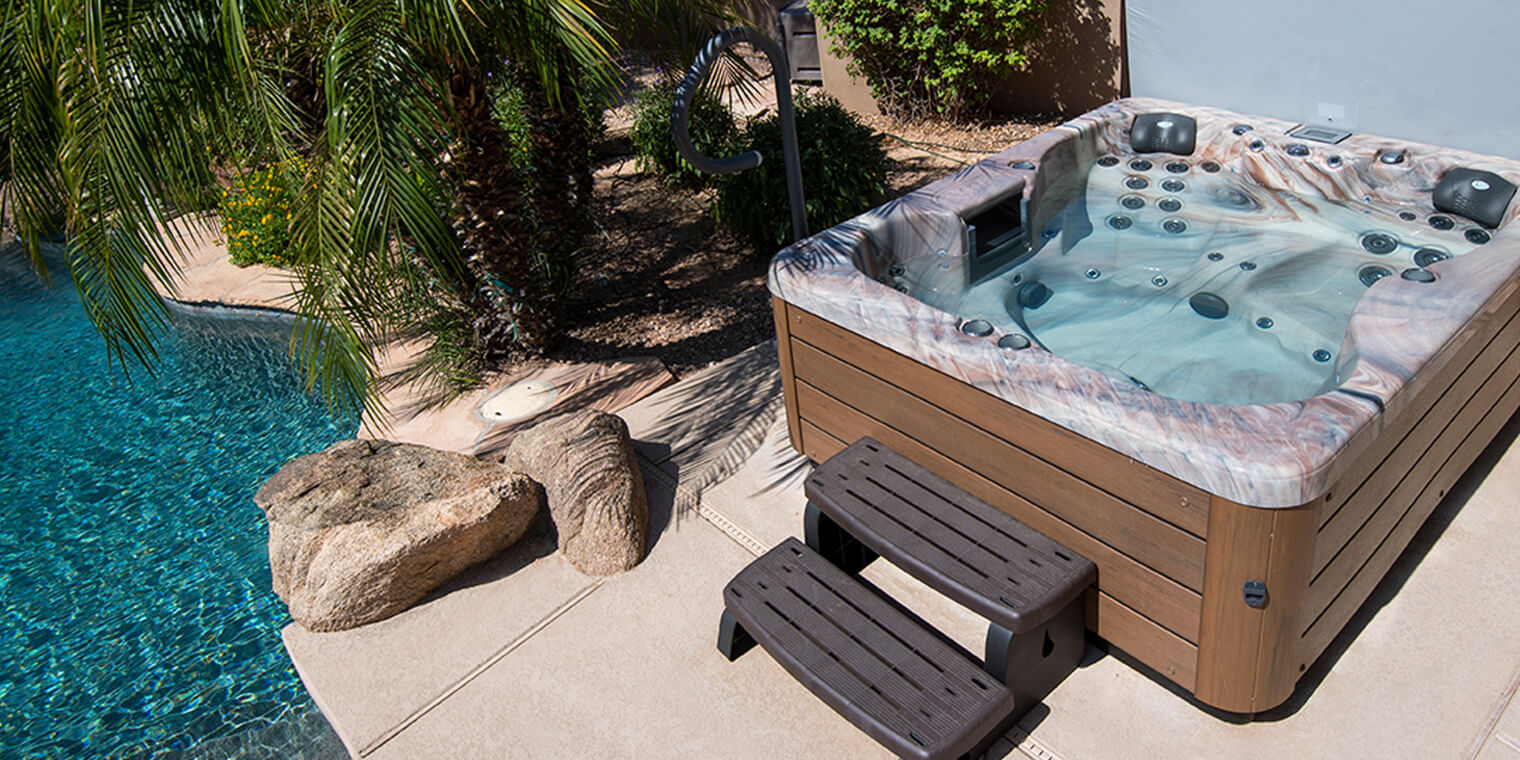 A Vita Spas hot tub with a wood-look cabinet on a stone patio beside a swimming pool, with steps up to the spa
